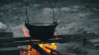 Kamp ateşinin üzerinde asılı duran siyah bir tencerenin yakın çekimi. Tencere kaynar su ve patatesle dolu..