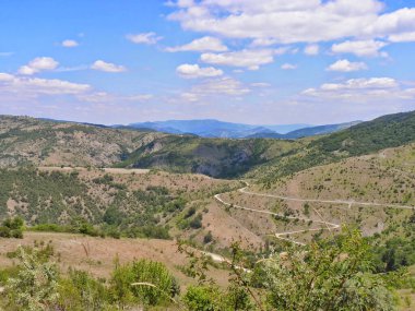 Görüntü, dağlık bir bölgeyi kesen dolambaçlı bir yolu olan manzaralı bir manzara tasvir ediyor. Yol, arazinin hatlarını takip eden bir dizi virajla döşenmiş bir otoyola benziyor.. 