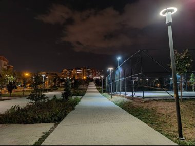 Gece sessiz bir şehir parkına giden yol aydınlandı. Parlak sokak lambaları ve basketbol sahasının sınırında. Gece gökyüzünün altındaki uzak hedeflere doğru bir yolculuğu simgeliyor..