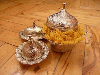 Zarif gümüş bir kase, sarmal makarnayla dolu, ahşap bir yüzeye yerleştirilmiş, süslemeli sadelikle mutfak sadeliğini birleştiriyor. Birbirine karışması gelenekler arasındaki uyumu yansıtır..