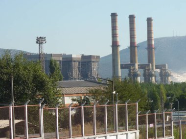 Yıpranmış Duman Yığınları Bir Endüstriyel Kompleksi Üzerine Yükleniyor: Açık Mavi Gökyüzü Altında Sanayi ve Doğanın Sessiz Bir Kavşağı, Çerçevelenmiş Yeşil ve Akdeniz 'de Zincirleme Çitli Çit