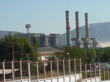 Yıpranmış Duman Yığınları Bir Endüstriyel Kompleksi Üzerine Yükleniyor: Açık Mavi Gökyüzü Altında Sanayi ve Doğanın Sessiz Bir Kavşağı, Çerçevelenmiş Yeşil ve Akdeniz 'de Zincirleme Çitli Çit