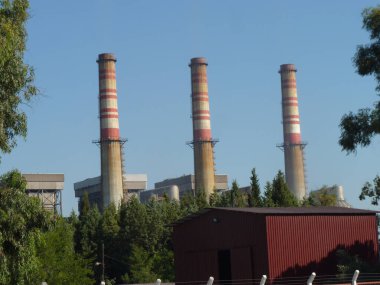 Weathered Smokestacks Towering Over an Industrial Complex: A Quiet Intersection of Industry and Nature Under a Clear Blue Sky, Framed by Greenery and a Chain-Link Fence in a Mediterranean Setting clipart