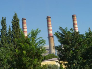 Yıpranmış Duman Yığınları Bir Endüstriyel Kompleksi Üzerine Yükleniyor: Açık Mavi Gökyüzü Altında Sanayi ve Doğanın Sessiz Bir Kavşağı, Çerçevelenmiş Yeşil ve Akdeniz 'de Zincirleme Çitli Çit