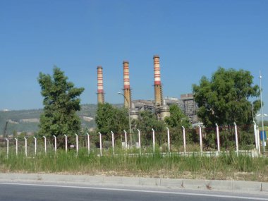 Yıpranmış Duman Yığınları Bir Endüstriyel Kompleksi Üzerine Yükleniyor: Açık Mavi Gökyüzü Altında Sanayi ve Doğanın Sessiz Bir Kavşağı, Çerçevelenmiş Yeşil ve Akdeniz 'de Zincirleme Çitli Çit