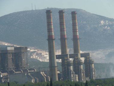 Yıpranmış Duman Yığınları Bir Endüstriyel Kompleksi Üzerine Yükleniyor: Açık Mavi Gökyüzü Altında Sanayi ve Doğanın Sessiz Bir Kavşağı, Çerçevelenmiş Yeşil ve Akdeniz 'de Zincirleme Çitli Çit