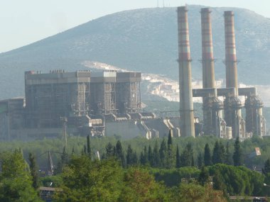 Yıpranmış Duman Yığınları Bir Endüstriyel Kompleksi Üzerine Yükleniyor: Açık Mavi Gökyüzü Altında Sanayi ve Doğanın Sessiz Bir Kavşağı, Çerçevelenmiş Yeşil ve Akdeniz 'de Zincirleme Çitli Çit
