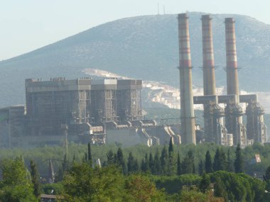 Yıpranmış Duman Yığınları Bir Endüstriyel Kompleksi Üzerine Yükleniyor: Açık Mavi Gökyüzü Altında Sanayi ve Doğanın Sessiz Bir Kavşağı, Çerçevelenmiş Yeşil ve Akdeniz 'de Zincirleme Çitli Çit