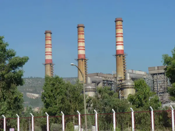 Yıpranmış Duman Yığınları Bir Endüstriyel Kompleksi Üzerine Yükleniyor: Açık Mavi Gökyüzü Altında Sanayi ve Doğanın Sessiz Bir Kavşağı, Çerçevelenmiş Yeşil ve Akdeniz 'de Zincirleme Çitli Çit