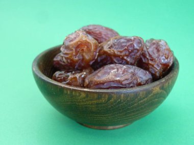 A wooden bowl filled with plump, glossy dates symbolizes abundance, nourishment, and the sweetness of life. Against a vibrant green background, these dates represent both natural simplicity. clipart