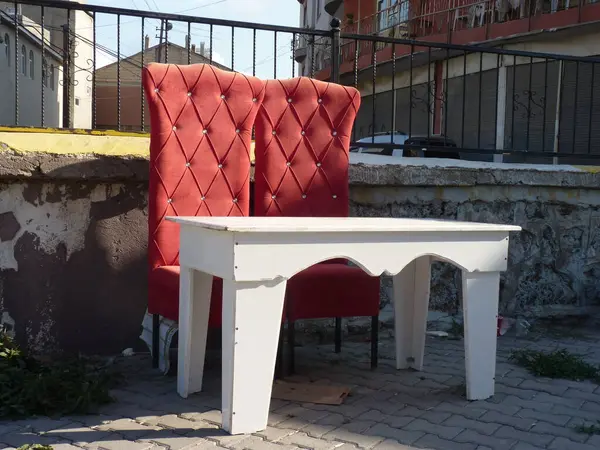 Beyaz masa ve kırmızı sandalyeli sıcacık bir teras sahnesi, basitliği, dengeyi ve doğa ile insan tasarımının samimi bir açık hava ortamında harmanlanmasını sembolize ediyor..