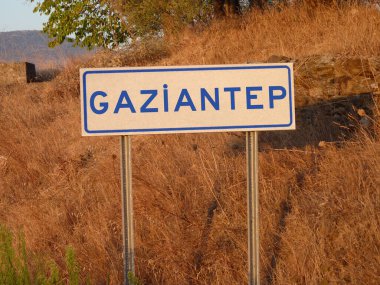 Blue 'Gaziantep' Sign on a Metal Pole: A Bold Contrast Against the Natural Landscape of Brown Grass and Soft Shadows, Capturing a Serene Afternoon in the Heart of the City. clipart