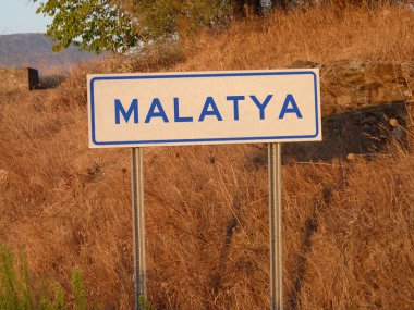 Blue 'Malatya' Sign on a Metal Pole: A Bold Contrast Against the Natural Landscape of Brown Grass and Soft Shadows, Capturing a Serene Afternoon in the Heart of the City. clipart