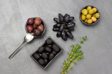 Green, red and black olives in bowls. Sprig of thyme and a bunch of black grapes. Copy space. Flat lay. Grey background.
