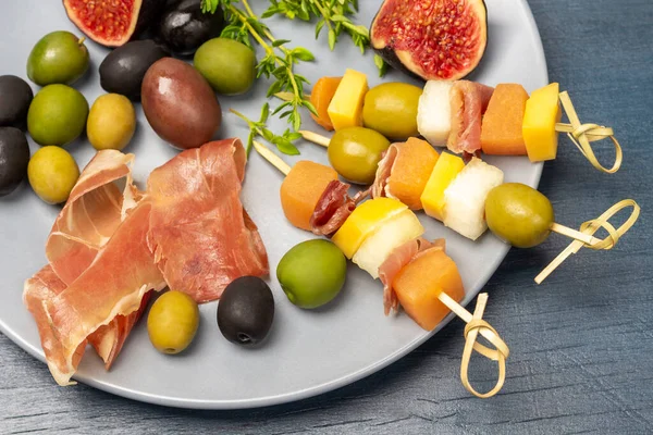 Skewers with olives, cheese, and melon on a gray plate. Ham, olives, fig halves and thyme sprigs. Flat lay. Blue background.