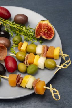 Cheese, olives, jamon on a skewer, Figs and sprigs of thyme on a gray plate. Top view. Blue background.