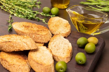 Olive oil in a glass bowl. Toasted bread and green olives on a board. Thyme sprigs. Brown background.