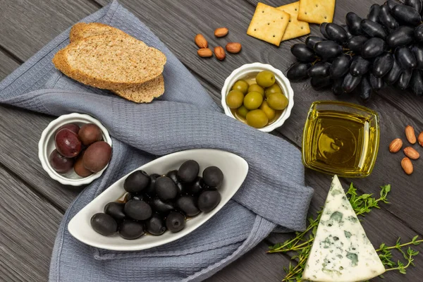 Black olives in a bowl and a bunch of black grapes. Blue cheese and nuts. Gray napkin. Flat lay. Dark wooden background.