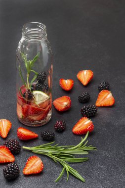Bir şişe dutlu su. Masada çilek ve böğürtlen var. Siyah arkaplan.
