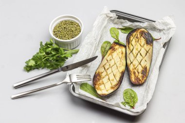 Grilled baked eggplants on pallet. Fork and knife. Mash in bowl and parsley sprigs. Gray background. Top view.