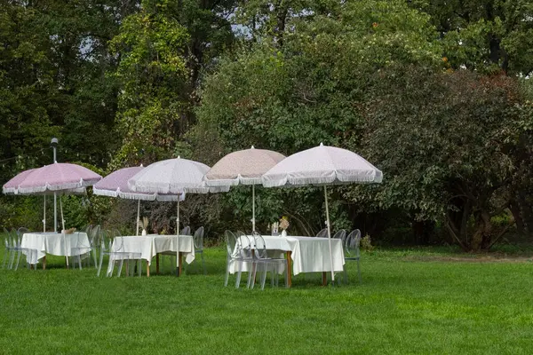 Bahçedeki yeşil çimlerin üzerinde beyaz masa örtüleri olan dört masa. Masaların yanındaki bahçe şemsiyeleri..