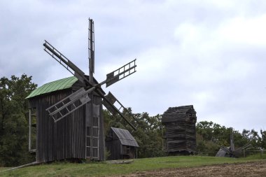 Açık hava parkındaki eski ahşap değirmenler. Çimenler yerde, ormanlar arkada. Bulutlu bir gün.