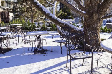 Büyük bir ağaç gövdesinin yanında bir kış kafesinin boş terası. Masalar ve metal sandalyeler karın altında..