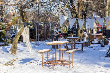 Gün batımında kış kafesi terasında karla kaplı masa ve banklar. Kardaki gölgeler. Kumaş peçeteler ağaçların arasında bir ipe asılı..
