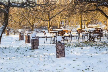 Parktaki ağaçların arasında karla kaplı masalar ve sandalyeler. Yerde kar var. Bir kış kafesinin açık terasında fenerler..