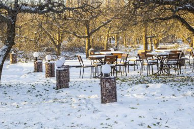 Parktaki ağaçların arasında karla kaplı masalar ve sandalyeler. Yerde kar var. Bir kış kafesinin açık terasında fenerler..