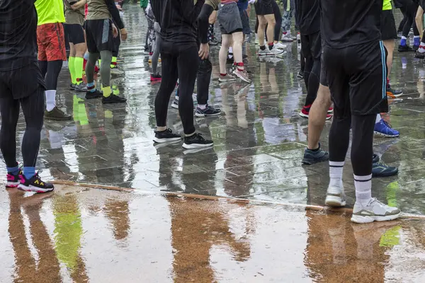 Sporcular yağmurda ıslak bir pistte dururlar. Su birikintilerindeki ayak yansımaları. Çerçevenin bir parçası