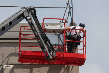 Başlık takan bir inşaat işçisi, bir inşaat alanında kırmızı kiraz toplayıcı kullanıyor, dış duvar kaplamasını onarıyor, yükseklikte güvenlik ve sanayi çalışmalarını vurguluyor.