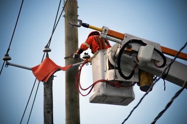 Yükseltilmiş bir çalışma platformundan elektrik hatlarında bakım yapan elektrik hattı çalışanı. Güvenli ve güvenilir elektrik hizmetleri sağlamak için gerekli. Mavi gökyüzünde turuncu yüksek görüş yeleği ve kablo tamir eden şapkası vardı..