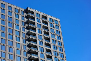 Açık mavi gökyüzüne karşı kurulmuş, balkonları olan, modern yüksek katlı beton binanın alçak açılı görüntüsü. Şehir mimarisini minimalist bir tasarım ile yakalar, simetri ve geometrik çizgileri vurgular.