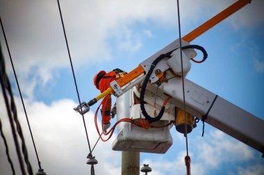 Elektrik hattı işçisi, yüksek bir çalışma platformundan elektrik hatlarını koruyarak güvenli ve güvenilir bir elektrik hizmeti sağlıyor. Turuncu, yüksek görüş mesafesi olan bir yelek ve şapka giyip açık mavi bir gökyüzünün altında kabloları onarmaya çalışıyor..