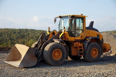 Yellow front loader parked at a construction site, equipped with a large bucket for earthmoving and material handling, set against a scenic background of trees and gravel. clipart