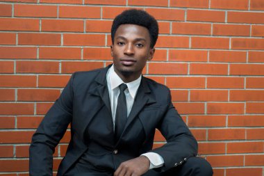 Siyah takım elbiseli ve kravatlı kendinden emin bir gencin portresi, tuğla bir duvara yaslanmış, profesyonellik, tarz ve modern, cilalı bir görünüm sergiliyor..