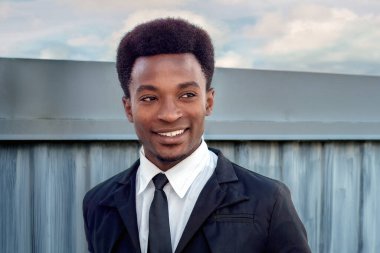 Siyah takım elbiseli ve kravatlı gülümseyen genç bir adamın portresi, açık havada metal bir çitin önünde duruyor, parlak gökyüzü arkaplanı, kendine güvenini ve tarzını gösteriyor..