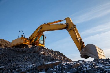 Kayalık bir inşaat sahasında çalışan sarı bir kazıcı, altyapı projelerinde ağır makineler taşıyor..