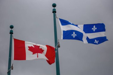 Kanada ve Quebec bayrakları gri gökyüzüne karşı birlikte dalgalanırken, ulusal ve taşralı gurur ve birliği sembolize ediyor.