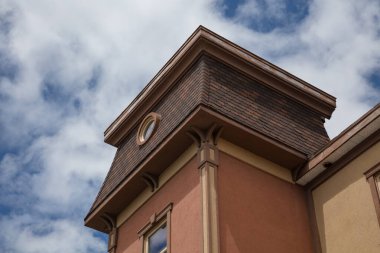 Yuvarlak pencereli, çatılı ve bulutlu mavi gökyüzüne karşı mimari detayları olan bir kuleye sahip klasik tarzda bir binanın köşesi. Emlak ve mimari temalar için mükemmel.