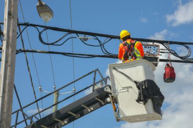 Güvenlik teçhizatı takan bir kamu görevlisi açık mavi gökyüzünün altındaki bir kova kaldırıcının elektrik kablolarını tamir ediyor. Enerji, altyapı ve güvenlikle ilgili görseller için mükemmel..