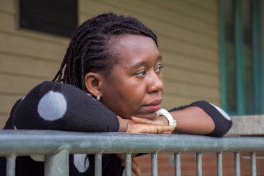 Benekli süveter giyen düşünceli Afrikalı kadın parmaklıklara yaslanıyor, elleri çenesinde, binanın dışındaki derin düşüncelere dalıyor. Yansıma, bekleme ya da farkındalık kavramları için idealdir.