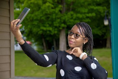 Güneşli yeşil bir parkta akıllı telefonuyla selfie çeken mutlu Afrikalı Amerikalı kadın. Yaz temaları, güzellik, eğlence, gezici yaşam tarzı ve tatil fotoğrafçılığı için mükemmel.