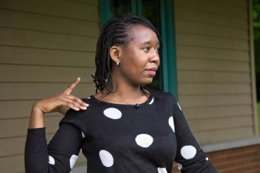 Örgülü saçlı, benekli kazak giyen Afrikalı bir kadın profil görüntüsünde el hareketi yapıyor ahşap duvarın önünde duruyor iletişim yaşam tarzı ve ifade teması için ideal.