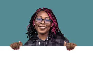 Gözlüklü, renkli saçlı ve elinde beyaz bir tabela tutan neşeli Afro-Amerikan kadın. Reklamlar, reklamlar, duyurular, pazarlama kampanyaları ve kişiselleştirilebilir mesajlar için mükemmel..