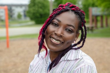 Mor örgülü, çizgili gömlek giyen, açık hava parkındaki kameraya güvenle gülümseyen mutlu genç siyahi kadın portresi.