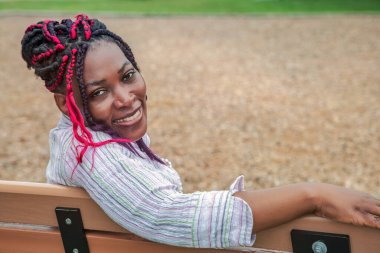 Çizgili tişörtlü mutlu siyah kadın doğal park ortamında ahşap bankta dinleniyor, eğlence ve yaşam tarzı kavramları için mükemmel.