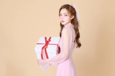 Genç, sevimli Asyalı bir kadın pembe zarif bir elbise içinde mutlu, Kore tarzı makyaj, nemlendirilmiş, pürüzsüz, bej arka planda hediye kutusu olan mükemmel bir ten. Yüz bakımı, kozmetoloji, estetik cerrahi..