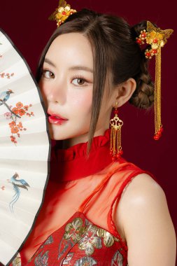 Temiz tenli güzel bir Asyalı kadın geleneksel Cheongsam Qipao elbisesi giyiyor ve elinde kırmızı arka planda poz veriyor. Stüdyoda kadın modelin portresi. Mutlu Çin Yeni Yılı.